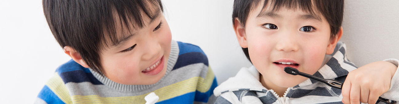 男の子二人が歯ブラシを持って座り、右側の男の子が歯を磨こうとするのを左側の男の子が見ている写真