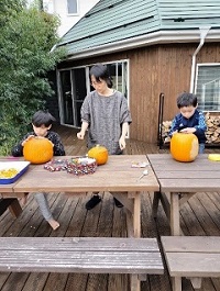 外に置かれた机上にオレンジ色のカボチャが置かれ2人の子供がそれぞれのカボチャで作品を作成している写真