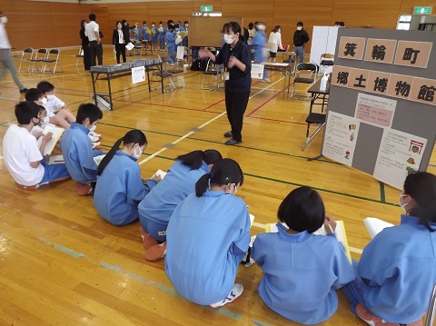 体育館内の箕輪町郷土博物館と書かれたボードの前に座った水色のジャージや体操服を着た10名の生徒たちが、向かい合って立つ女性の説明を聞いている写真