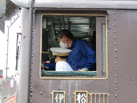 電気機関車ED19-1の運転席で、駅員の帽子を被った子供が青色のシャツを着た男性から運転操作を教えてもらっている様子を外から撮影した写真
