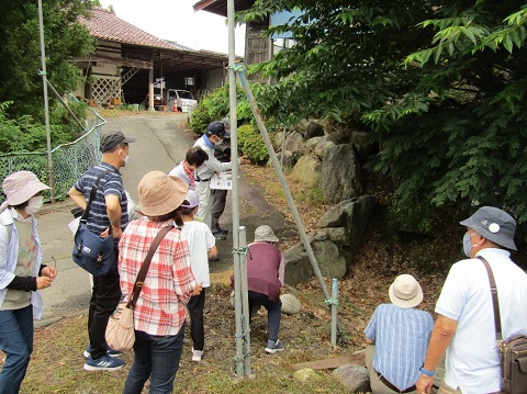 急勾配の坂沿いにある苔が生えた大きな石の前で、帽子を被った参加者たちが紺色のキャップを被った男性の話を熱心に聞いている写真
