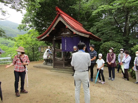 お地蔵様が祀られている赤色の屋根のお堂の前に立つ男性の話を、参加者たちが聞いている様子の写真