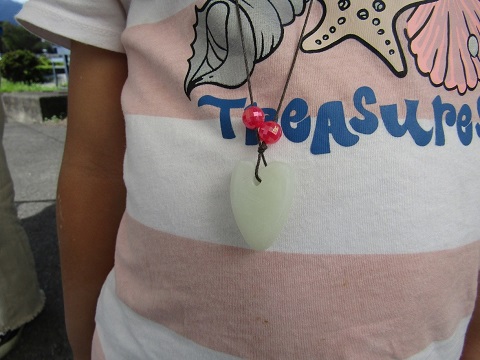 白とピンク色のボーダー柄のシャツを着た女の子が、ハート型に成形した石に2つの赤色のビーズと紐をとおして首にかけた様子をアップで撮影した写真
