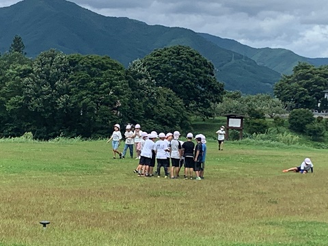 大きな山や周囲に木々が生えた自然豊かな芝生広場で、白色の帽子を被った児童たちが集合したり歩いたりしている写真