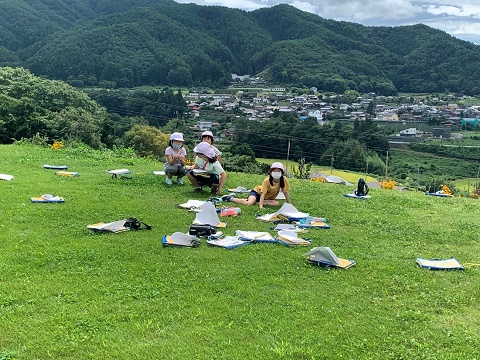 遠くに住宅が見渡せる高台の芝生広場で、4名の女子児童がシロツメクサを摘んでいる様子の写真