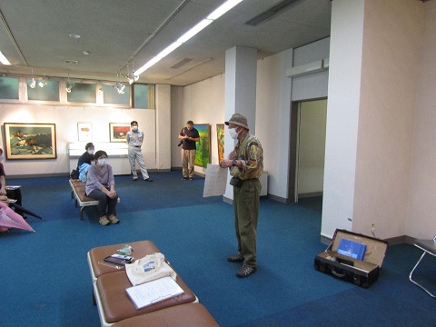 絵画作品が展示された館内で、前方に立つ帽子を被った男性講師の話を椅子に座った参加者が聞いている写真