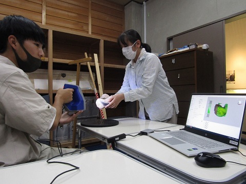 3Dスキャン装置を持った男子学生が、白シャツを着た女性が持った土偶の底をスキャンしながらパソコンの画面を見ている様子の写真