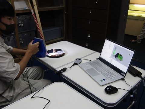 黒色の回転台の上に置かれた小さな土偶に3Dスキャン装置をあてた男子学生が、パソコンに映し出された画面を見ながらスキャンを行っている様子の写真