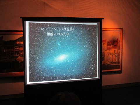 赤色の照明が照らされ薄暗くなった館内にスクリーンが設置され、アンドロメダ星雲が映し出されている写真
