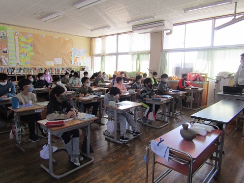前方に設置された机の上に土器の様な物が置かれ、教室の左前に立っている男性の話を、自席に座った児童たちが熱心に聞いている様子の写真