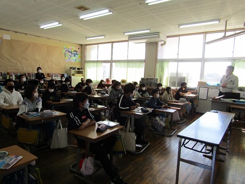 教室の左前に立っている男性の話を、自席に座った児童たちや教室の後ろに立っている先生が聞いている様子の写真