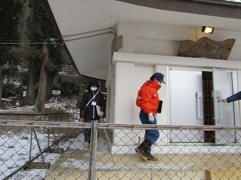 斜めに雪が降っている寒い中、防寒着を着た2名の担当者が、無量寺のスタッフが開けた文化財のある建物の扉の中に入って行こうとするパトロールの様子の写真