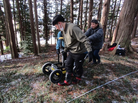 木々が生えている急勾配の古墳の上を、車輪がついた機械を3人がかりで動かし地中レーダー探査調査を行っている様子の写真