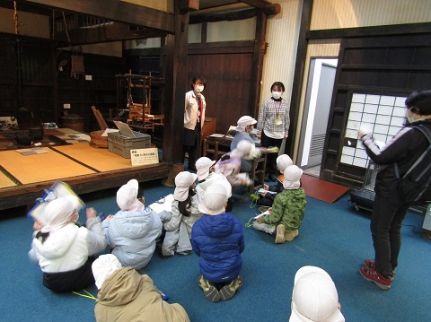 畳敷きの昔の住宅の中に囲炉裏などの道具が設置された館内で、女性スタッフから説明を受けている白色の帽子を被った児童たちの後ろ姿の写真