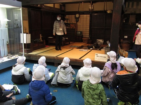 畳敷きの昔の住宅の中に設置された囲炉裏の傍に女性スタッフが立って説明を行っているのを、白色の帽子を被った児童たちが熱心に聞いている後ろ姿の写真
