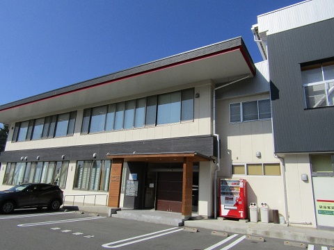入り口前に駐車場が設けられた、ベージュ色と黒色の外壁で2階建ての仮事務所となる産業支援センターみのわの建物外観の写真