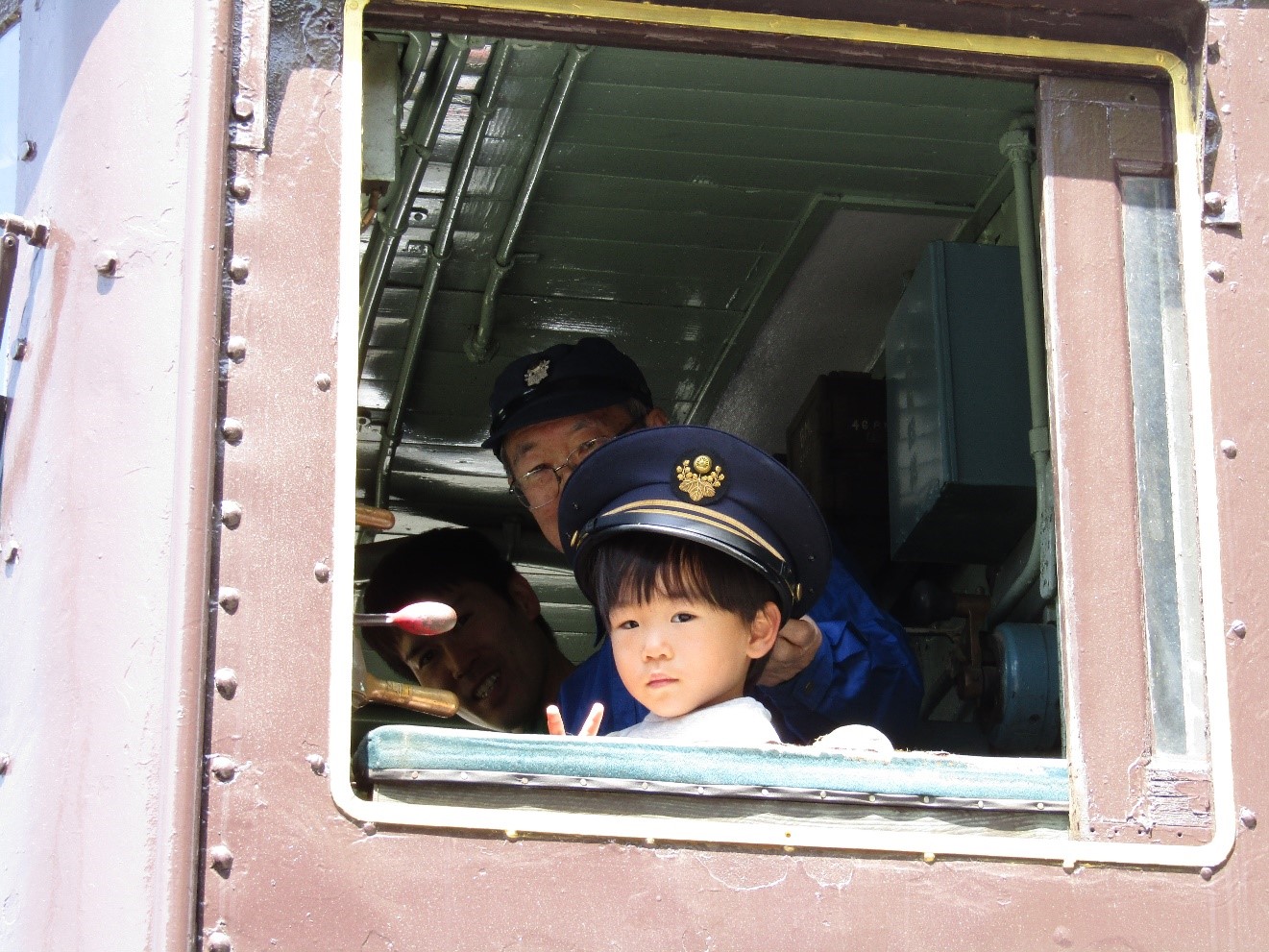 電気機関車の運転席の窓から車掌の帽子をかぶり、ピースをしてこちらを見る男の子
