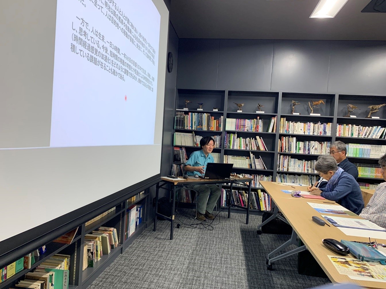 スクリーンの横で話をする講師と話しを聞きながらメモを取る聴講者