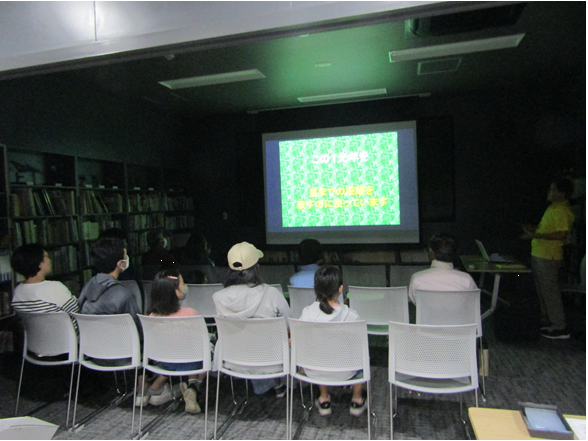 博物館の図書・講座室でスクリーンを見ながら、野口先生の話を聞く親子