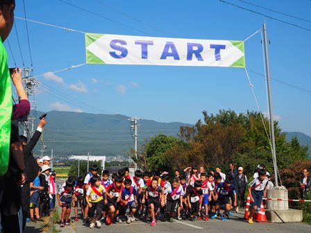 左側に立っている男性が右手に持って上げたスターターピストルの合図と同時に、肩に赤色のタスキを掛けた男子の選手が一斉に走り出した様子の写真