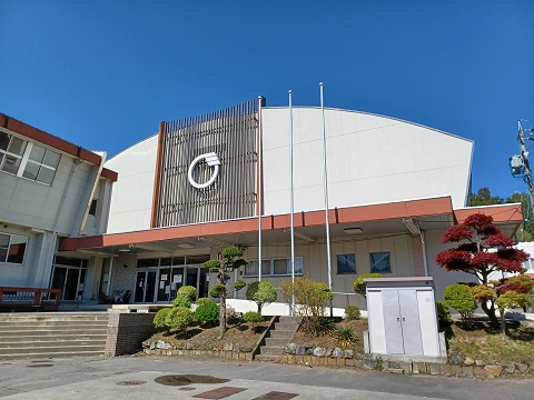 湾曲した屋根で、白壁の外壁に縦縞の格子のようなデザインに町章が施された箕輪町町民体育館の建物外観の写真