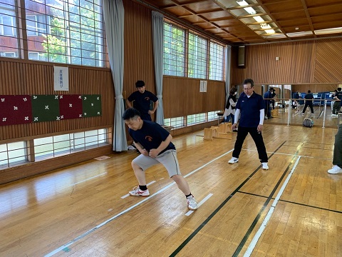 体育館の屋内で、2本の白いラインに両足を乗せた男性が反復横跳びを行っている様子の写真