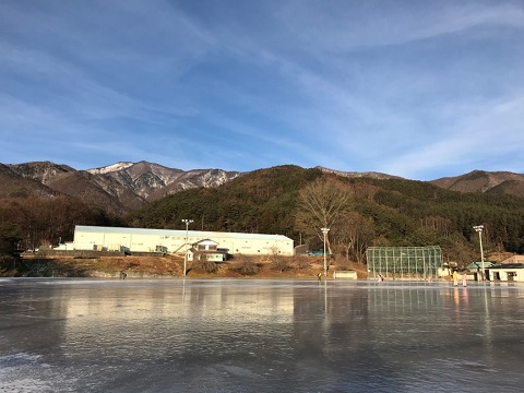 薄っすら雪が積もった山並みの麓に、ナイター照明が設置された多目的運動場の地面に氷が張られている上古田スケート場の写真