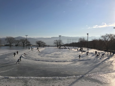 遠くに山脈が見渡せる地面に氷が張られた多目的運動場で、スケートを楽しんでいる人達の写真