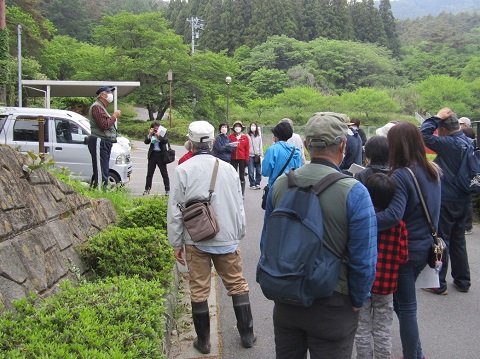 山や森などの自然に囲まれた細い道路上で、キャップを被った男性の話を参加者が聞いている写真