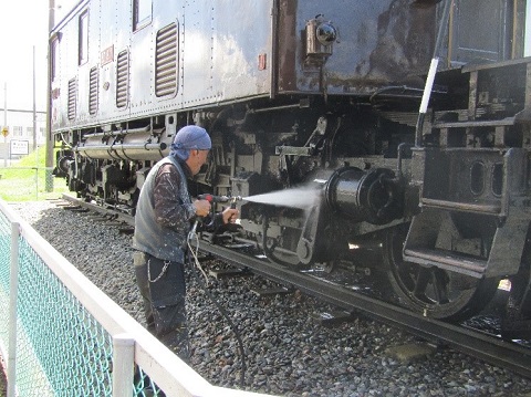 青色のタオルを頭に巻いた男性が、高圧洗浄機で電気機関車ED-19の車輪がついた下部分を洗浄している様子の写真