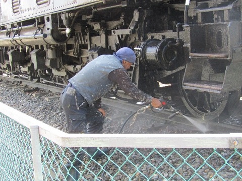 青色のタオルを頭に巻いた男性が、電気機関車ED-19が展示されている線路を右手に持った高圧洗浄機で洗浄している様子の写真