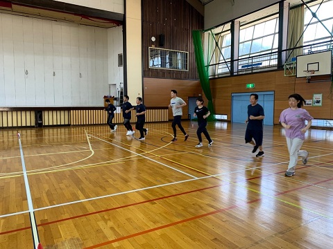 奥にステージがある体育館内で、横一列に並んだ7名の参加者が一斉に走り出している体力測定会の様子の写真