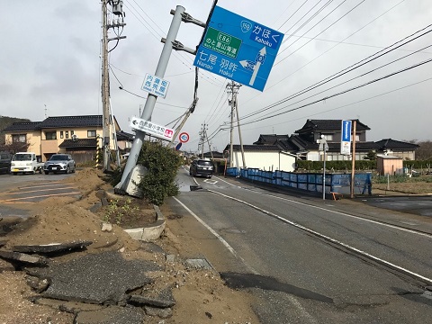 道路横の地面がめくれ上がり、青い道路標識が大きく傾いている写真