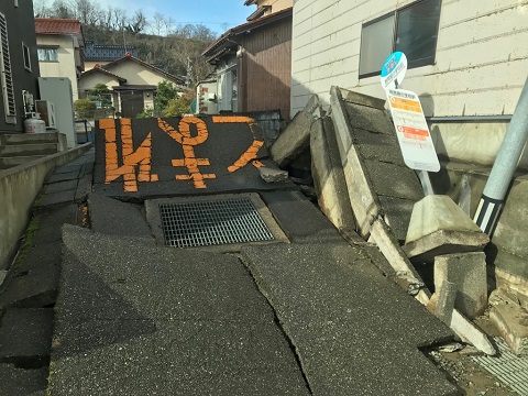 道路と道路横の側溝が大きく盛り上がり、バス停留所の標識版が倒れている写真
