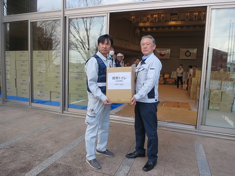 男性二人が「携帯トイレ」と書かれた紙が貼られているダンボールを一緒に持っている写真