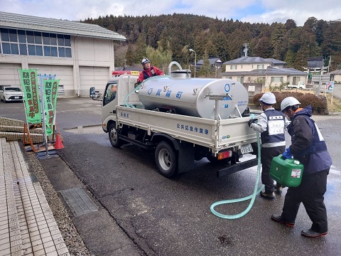 給水車のタンクの前方で男性が1人、給水車の後方で1人の方がホース、もう一人の方が緑の小さなタンクを持ち作業をしている様子の写真
