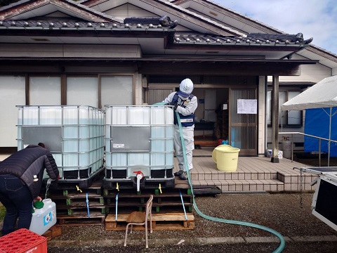 公民館のような建物の玄関先に大きなタンクが2つ置かれ、その中に作業員の方がホースで水を入れている様子の写真