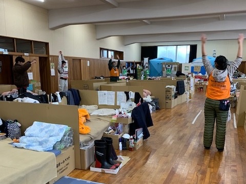 ダンボールベッドなどが置かれている避難所で、7名ほどの人が等間隔に広がって両腕を上に伸ばしストレッチをしている様子の写真
