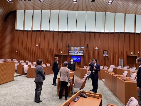 議会室の中央に設置された席の周りに4名の議員が立ち、右側に立っている男性の話を聞いている視察の様子の写真