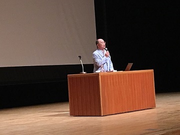 ステージの壇上に立ち、マイクを使って話をする田口一博先生の写真