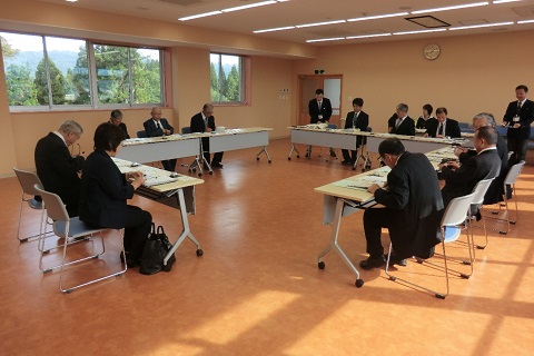 ロの字に設置された長机の席に議員の方々や関係者が座り、左奥の席から立ち上がった男性の話を机の上に置いた資料を見ながら聞いている視察研修の様子の写真