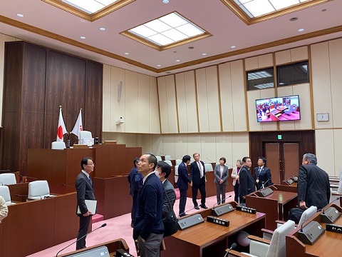 議会室内で議員の方々が集まっている様子が、左壁に設置されたモニターに映し出されている視察の様子の写真