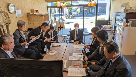 資料が置かれた6つの机が設置された室内で、机を囲んで議員の方々が座っている視察の様子の写真