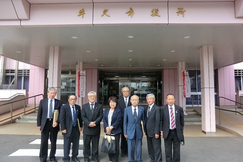 薄ピンク色の外壁の大きな軒先に「牛久市役所」と書かれた建物の前で、8名の議員の方々が並んでいる写真