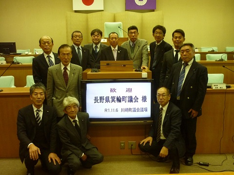 議会室の中央に設置された「歓迎 長野県箕輪町議会様 令和1年11月6日川崎町議会議場」と書かれたモニターを囲んで12名の議員の方々が集まっている写真