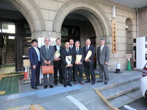 縦書きで会津若松市役所と書かれた看板が設置された建物の前で、8名の議員の方々が並んでいる視察研修の様子の写真