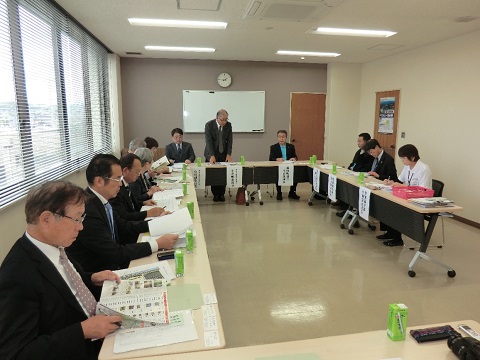 縦長の室内に設置された長机の席に議員の方々や関係者が座り、前方中央の席から立ち上がった男性の話を資料を見ながら聞いている視察研修の様子の写真