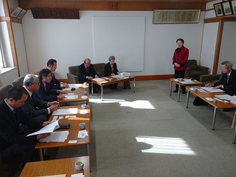 議員の方々や関係者が横長のテーブルに2名ずつ着席し、右前方の席から立ち上がった赤色の服を着た女性の話を聞いている視察研修の様子の写真