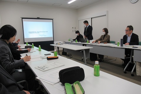 室内前方に資料が映し出されたスクリーンがあり、向かい合って設置された長机の席に議員の方々や関係者が座り、右側の2番目の席から立ち上がった男性の話を資料を見ながら聞いている視察研修の様子の写真