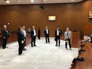 茶色の壁にモニターが設置された議会室内に立つ7名の議員の方々が、議会席に向かい合って立つ人の話を聞いている写真